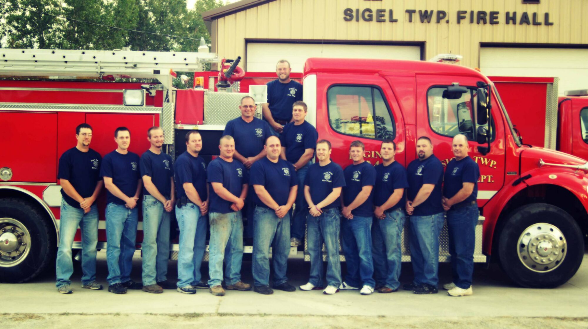 Group of firefighters posing with fire truck outside hall.